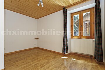 An empty room features wooden floor and ceiling, white walls, and a large window with plaid curtains and a radiator below it.