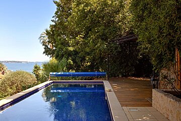 An outdoor swimming pool surrounded by a wooden deck and lush green trees overlooks the sea under a clear blue sky.