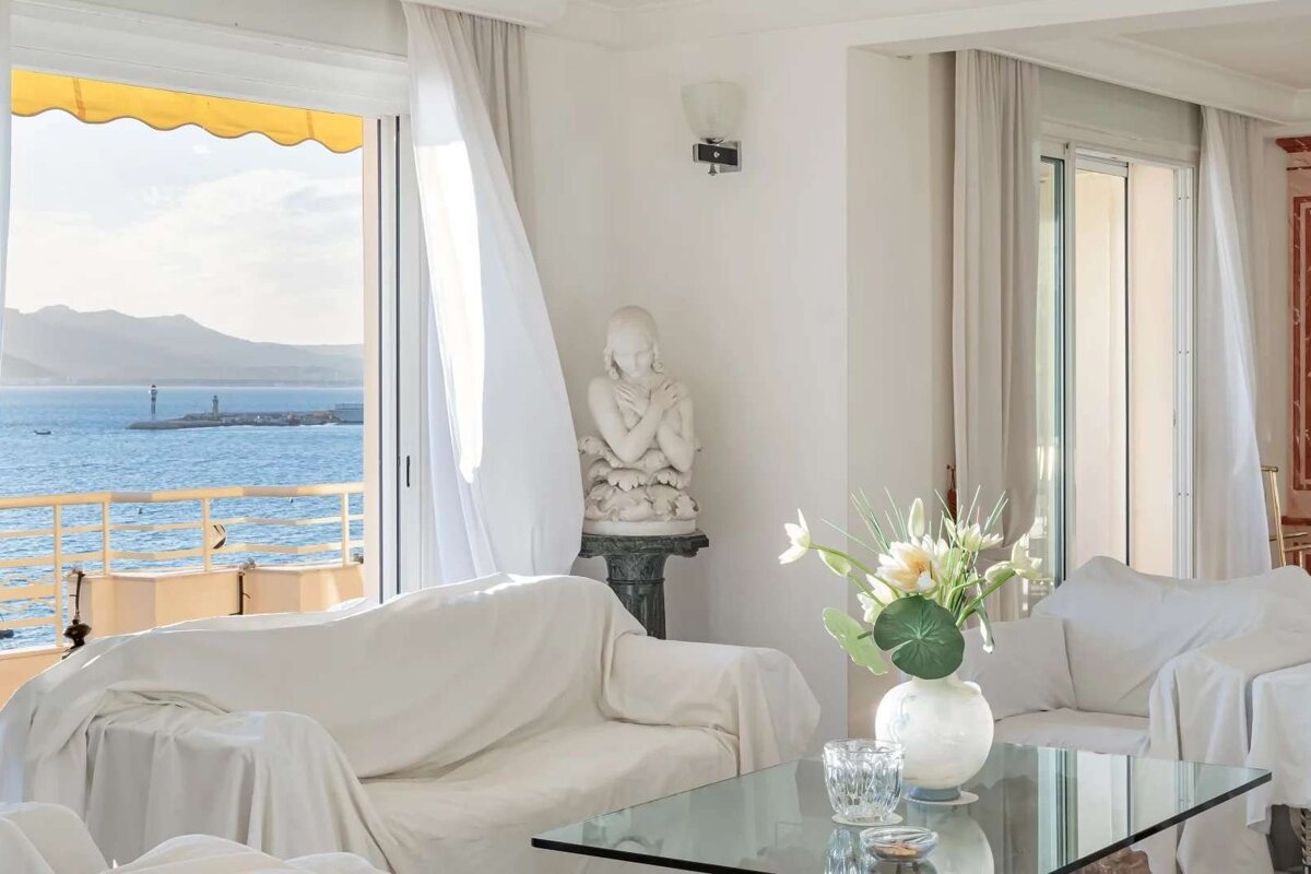 Bright white living room overlooking the sea, with white slipcovered furniture, a classical statue, and sheer curtains blowing in the breeze.