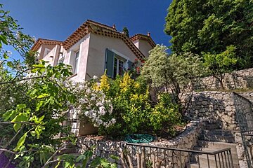A picturesque house with green shutters, surrounded by vibrant greenery, colorful flowers, and stone terraced steps under a clear blue sky.