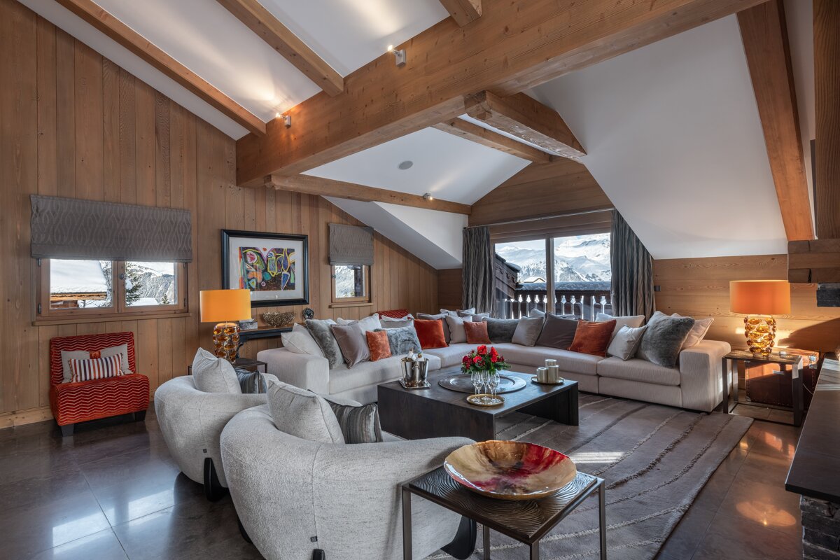 Cozy, luxurious chalet living room featuring wood-paneled walls, exposed beams, plush seating, and panoramic mountain views through large windows.