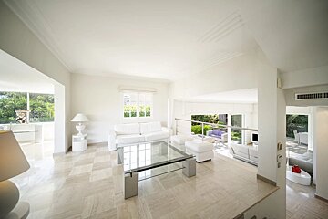 A spacious, modern living room featuring white furniture, a glass coffee table, beige tile flooring, and large windows.