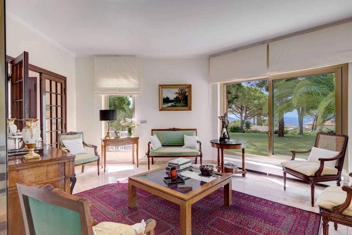 Elegant living room with traditional furniture, a red rug, and large windows revealing a lush garden and distant sea view.