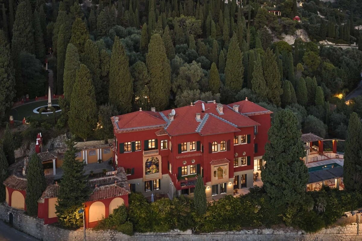 Aerial view of a grand red mansion with red roofs, nestled among dense, tall green trees on a hillside, featuring gardens and a pool.