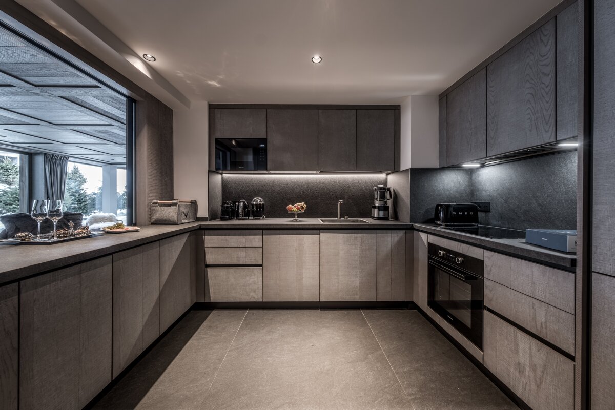 A modern, U-shaped kitchen with dark grey wood grain cabinets, sleek integrated appliances, under-cabinet lighting, and a large window to the outside.