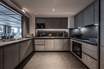 A modern grey and wood-toned U-shaped kitchen with under-cabinet lighting, contemporary appliances, and a large window overlooking an outdoor scene.