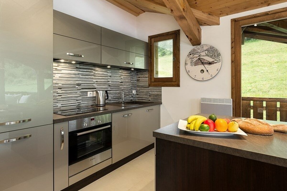 A modern kitchen featuring glossy grey cabinets, wooden ceiling beams, and a dark island with a bowl of fresh fruit.