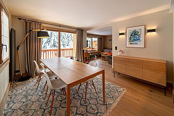 Modern dining room with a wooden table, stylish rug, and large windows revealing a snowy mountain view. It connects to a cozy living area.