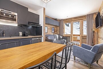 Open-plan mountain apartment with a sleek kitchen, wooden dining table, comfy living space, and stunning snowy views from the windows.