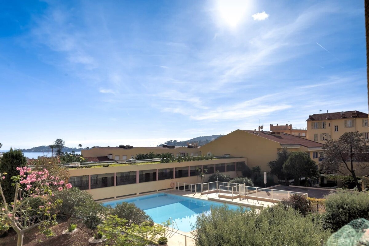 Sunny view of a blue outdoor swimming pool bordered by buildings, green foliage, and the sea in the distance under a clear sky.