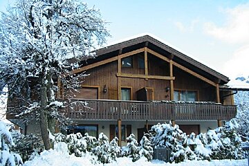 A cozy wooden chalet covered in snow, surrounded by snow-laden trees and bushes, under a bright winter sky.