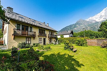 Sunny alpine scene: a yellow house with a garden, backed by towering mountains and clear blue skies.