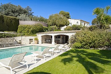 A beautiful sunny backyard featuring a swimming pool with lounge chairs, a stone cabana, and a green lawn under a clear blue sky.