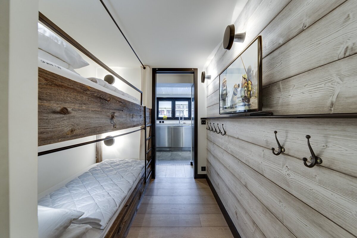 A modern rustic hallway features wooden bunk beds on the left, and a wood-paneled wall with a TV and coat hooks on the right, leading to a kitchen.
