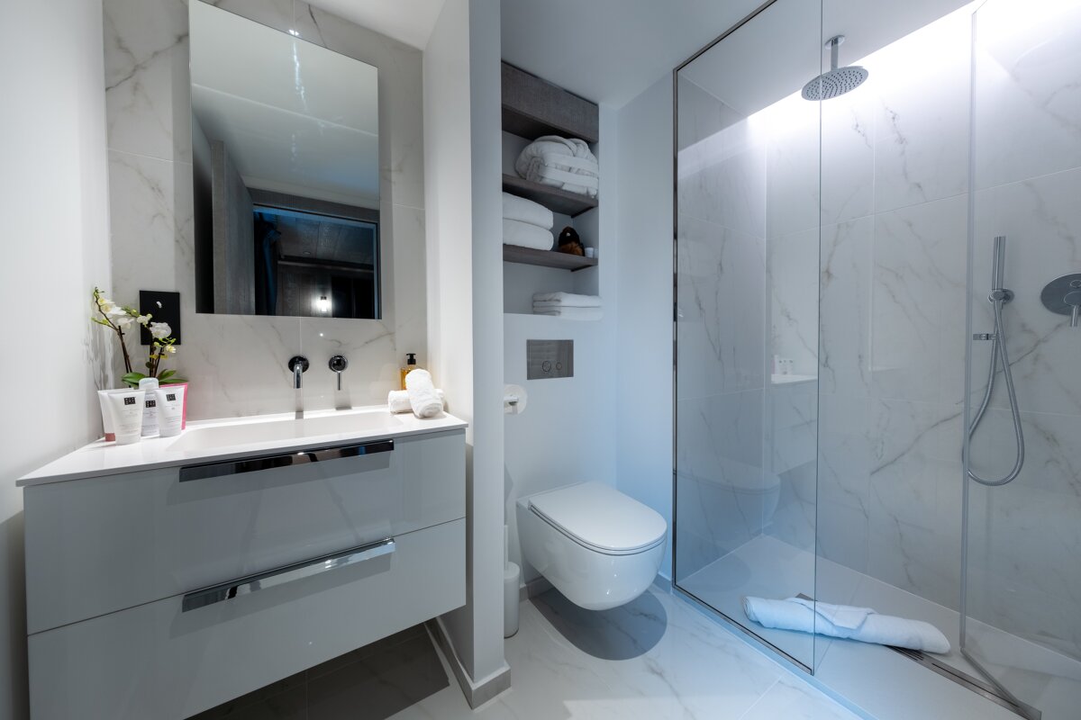 A modern, minimalist white bathroom with marble-look tiles, vanity, mirror, wall-mounted toilet, and a sleek glass shower.