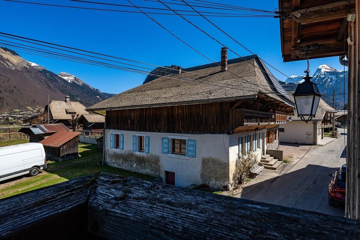 A traditional village house with a wooden roof and blue shutters, viewed from above, with snow-capped mountains and a clear blue sky in the background.