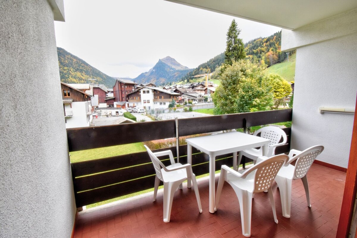 A balcony with white plastic table and chairs, offering a scenic view of a mountain village, forested hills, and a distant peak under an overcast sky.