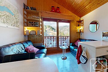 Cozy, wood-paneled room with black sofas, a kitchen counter, and a balcony offering a stunning view of a wooded mountain landscape.