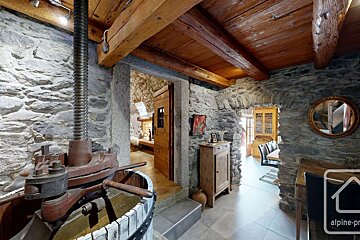 Rustic interior with stone walls, wooden beams, and an antique press dominating the foreground. Doorways reveal more cozy, traditional rooms.