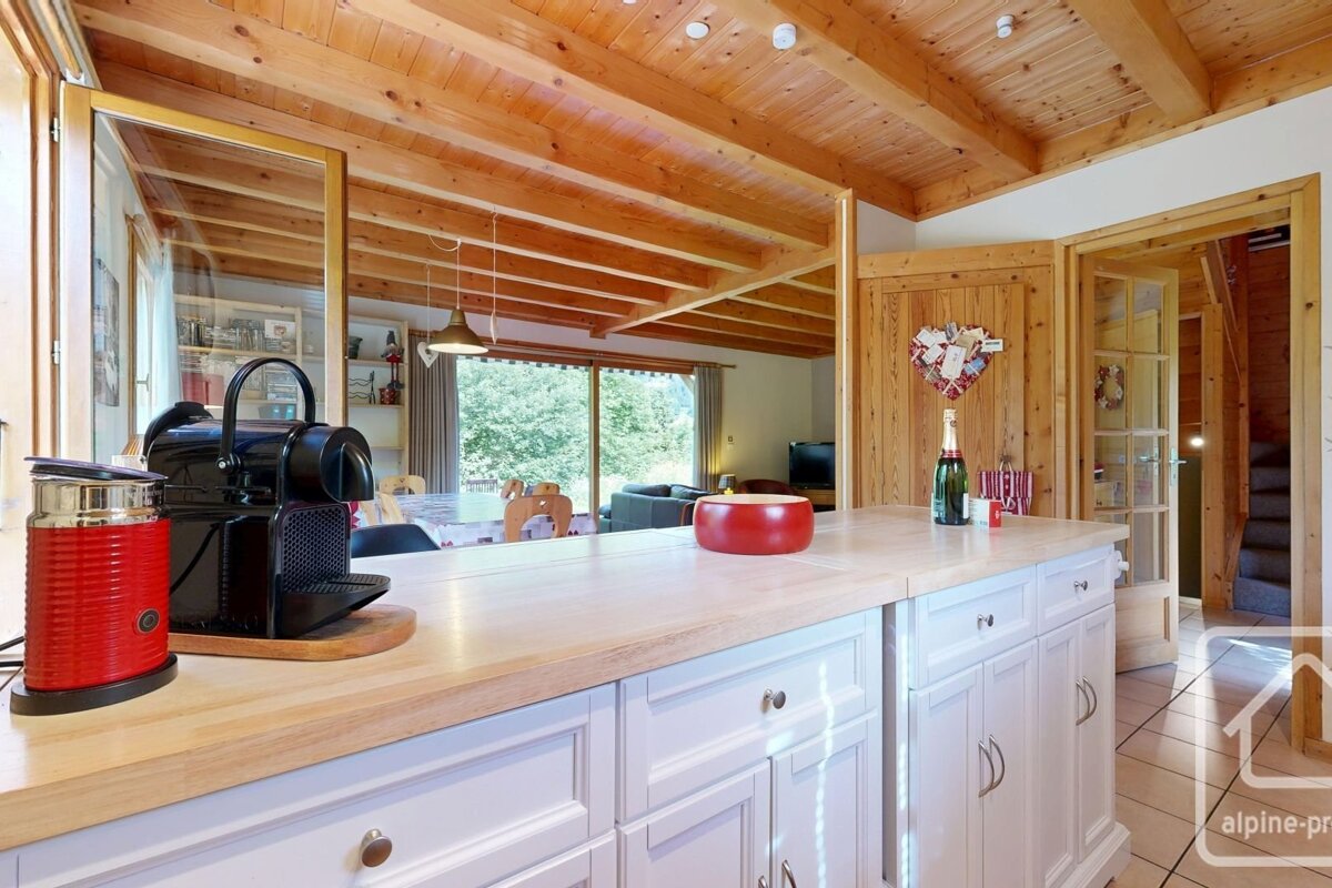 Cozy kitchen with wooden ceiling and white island featuring a coffee machine, champagne, and a view to a living room and trees outside.