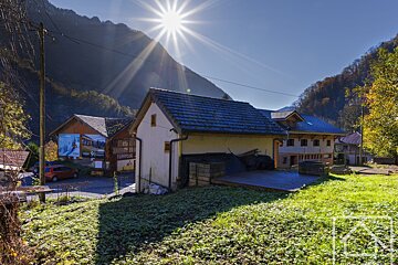 Sunny mountain village scene with traditional buildings, lush green grass, and a bright sunburst overhead, casting long shadows.