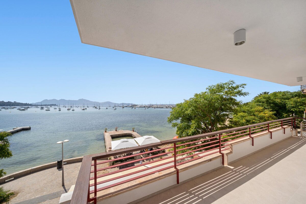 A bright balcony overlooks a bay filled with sailboats, distant mountains, and trees under a clear blue sky, with a pier and shaded area below.