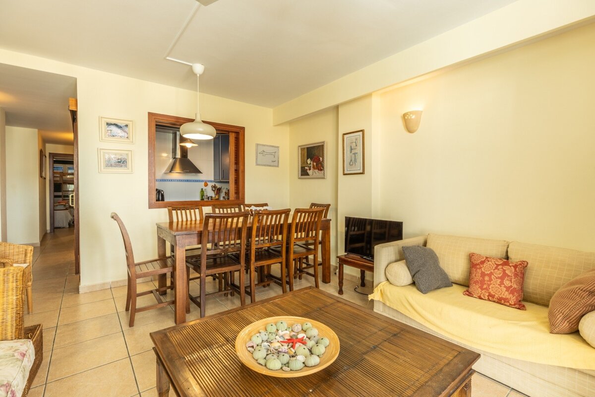 A cozy living/dining room with yellow walls, wooden furniture, a sofa, and a coffee table. A kitchen is visible through a pass-through window.