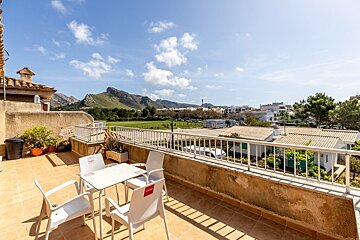 A tiled balcony with a white table and chairs overlooking a green landscape and mountains under a bright blue sky.