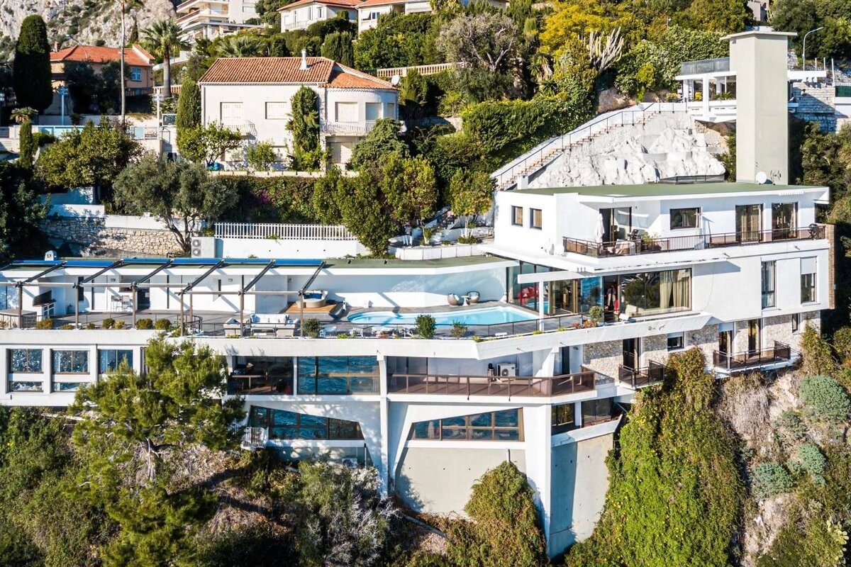 A modern, multi-level white mansion with a pool and large terraces, nestled into a green hillside amidst other homes and trees.