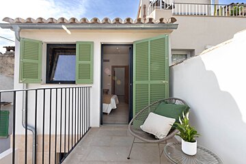 A bright balcony with open green shutters, a comfy chair, and a plant on a glass table. It offers a peek into a well-lit room beyond.