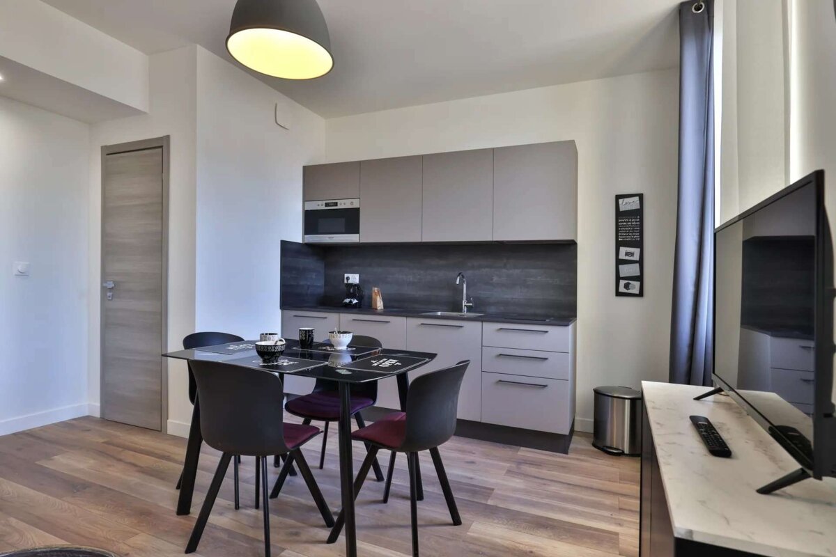 A modern, compact studio apartment featuring a grey kitchen, dining table with dark chairs, and a TV area, all on a wood-look floor.