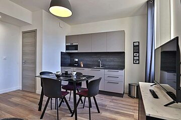 A modern, compact studio apartment featuring a grey kitchen, dining table with dark chairs, and a TV area, all on a wood-look floor.