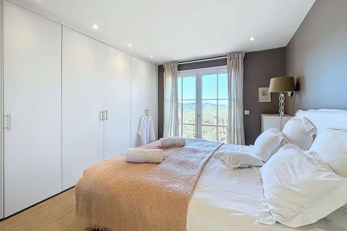 A bright bedroom with a large bed, white built-in wardrobes, a bedside lamp, and a window overlooking a scenic view.