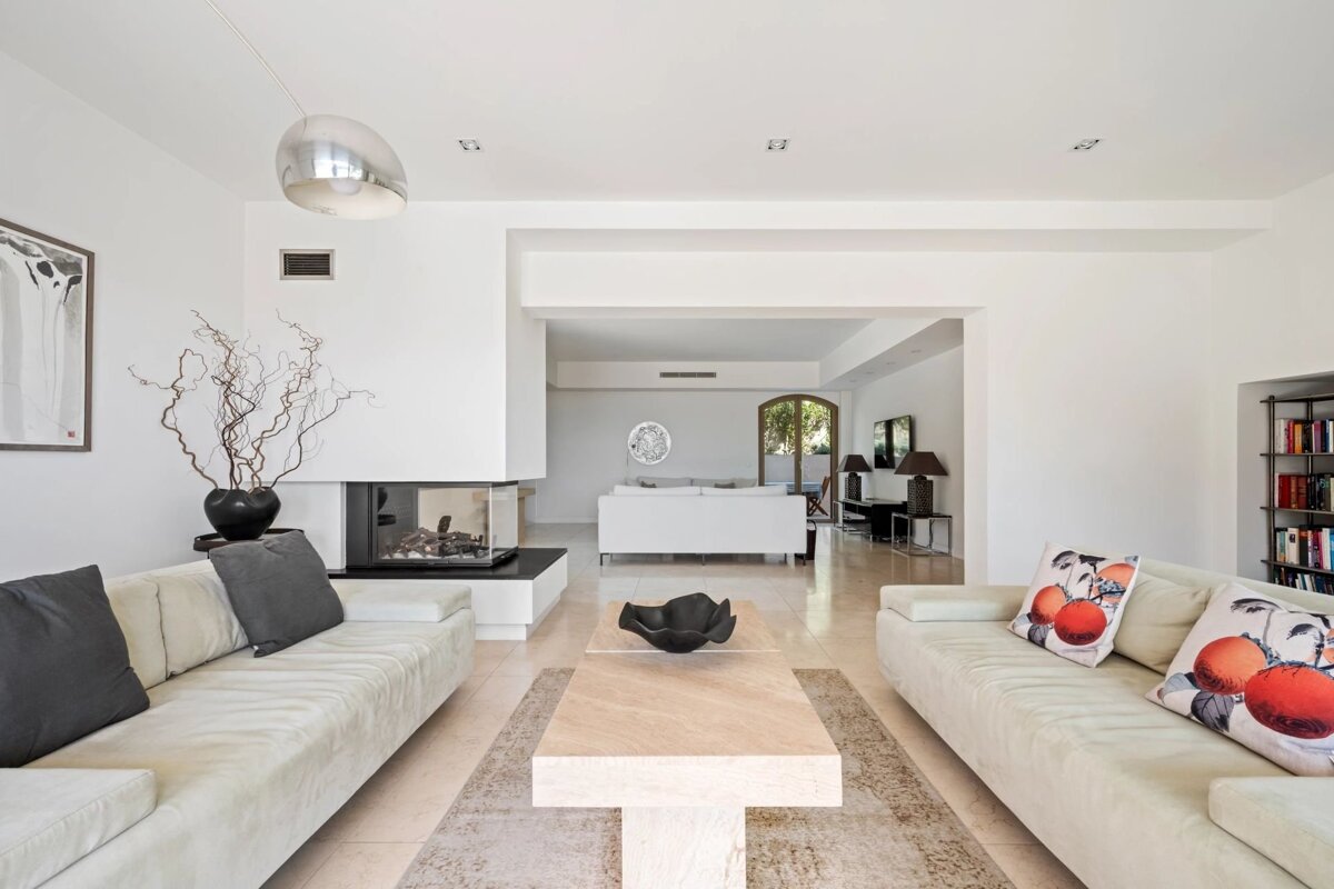 A bright, modern living room featuring two light sofas, a beige coffee table, and a sleek glass fireplace. The decor is minimalist with white walls.