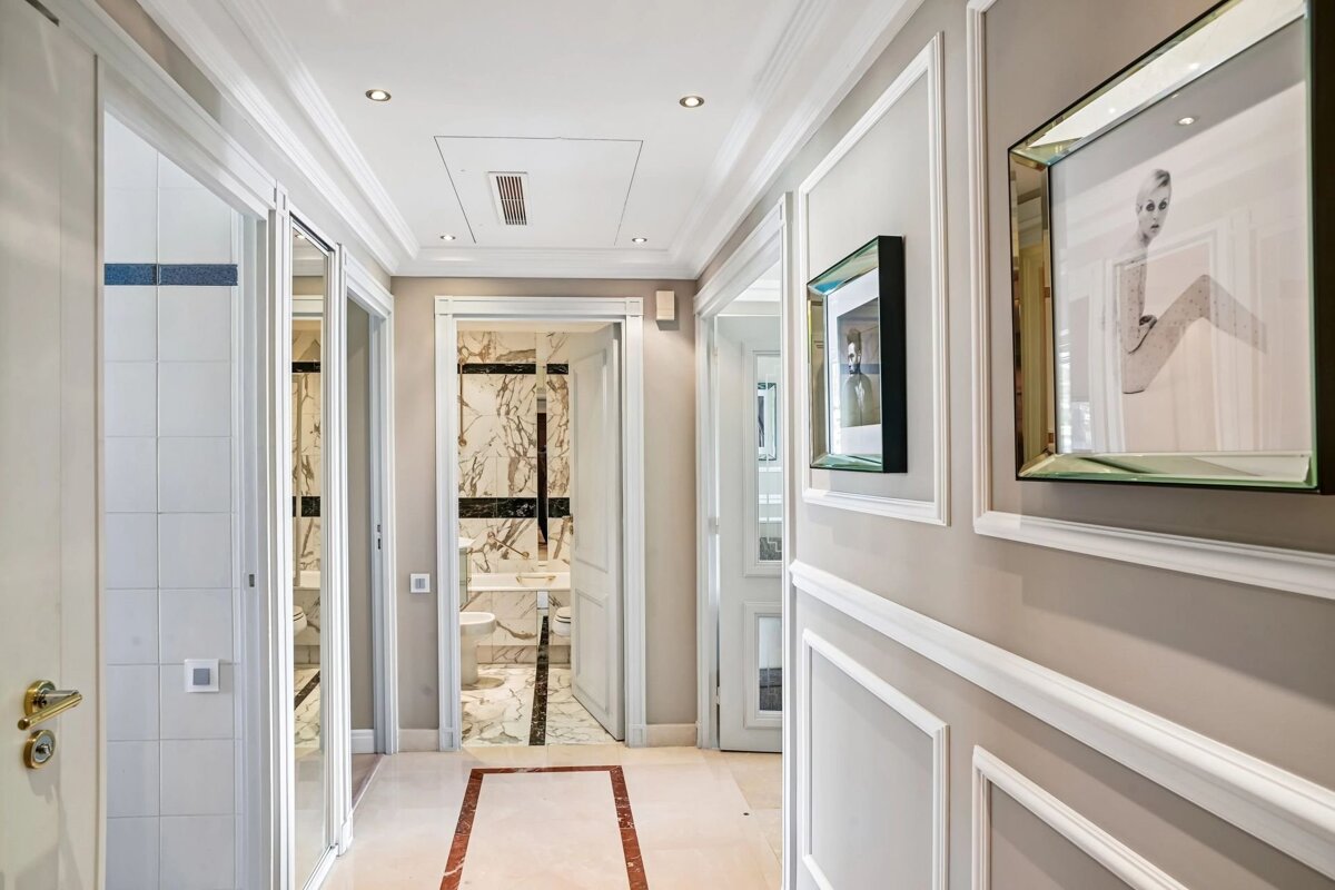 A well-lit, modern apartment hallway with beige walls, white wainscoting, framed art, and an open door to a marble bathroom.