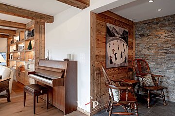 Cozy chalet interior with an upright piano, rustic wooden beams, and a seating area featuring stone and wood walls.