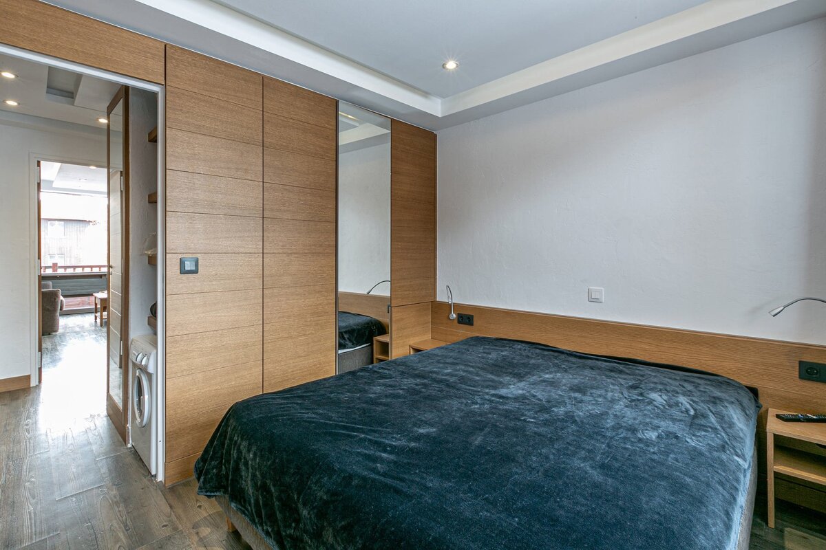 A modern bedroom with a large bed, wooden headboard, integrated wardrobe with a mirror, and a washing machine visible in a partially open closet.