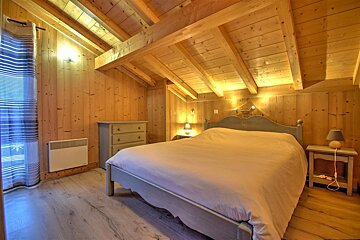 A cozy, rustic bedroom features light wood paneling, exposed beams, a large bed, dresser, and warm lighting, creating a chalet feel.