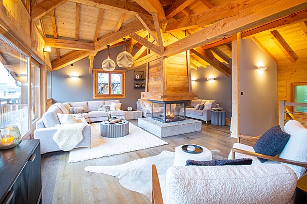 A cozy, modern chalet living room with exposed wooden beams, a central glass fireplace, plush sofas, and elegant decor, exuding warmth and comfort.
