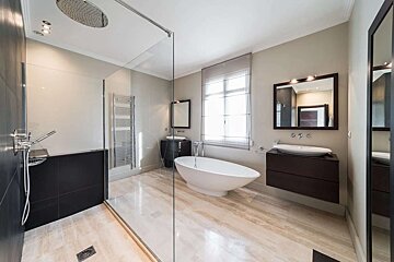 A sleek, modern bathroom with a glass shower, white freestanding tub, two dark wood vanities, and polished light marble floors.