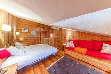 Cozy, wood-paneled attic room with a bed, red daybed, and a slanted ceiling, creating a warm, cabin-like atmosphere.