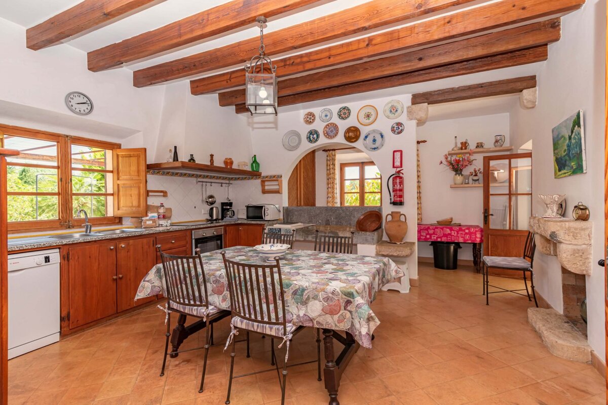 A rustic kitchen with exposed wooden beams, terracotta tiles, and a dining table. Features large windows, decorative plates, and an arched entry.