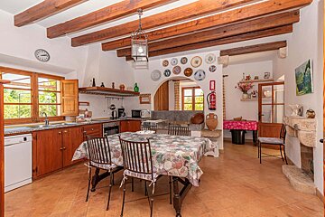 A rustic kitchen with exposed wooden beams, terracotta tiles, and a dining table. Features large windows, decorative plates, and an arched entry.