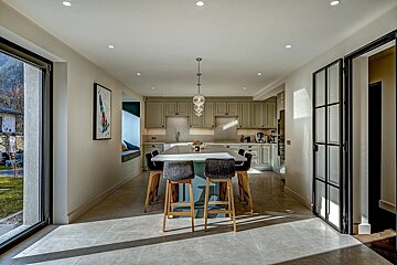 A bright, modern kitchen features sage green cabinets, a large island with bar stools, and natural light streaming through glass doors, casting long shadows.