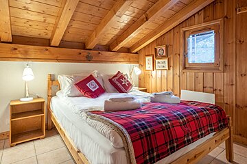 Cozy, rustic bedroom with a wooden slanted ceiling, paneled walls, a bed featuring a red plaid blanket, accent pillows, and a small window.