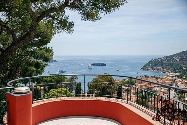 A stunning balcony view overlooks a deep blue bay with cruise ships and yachts, a charming coastal town with red roofs, and lush green hills.