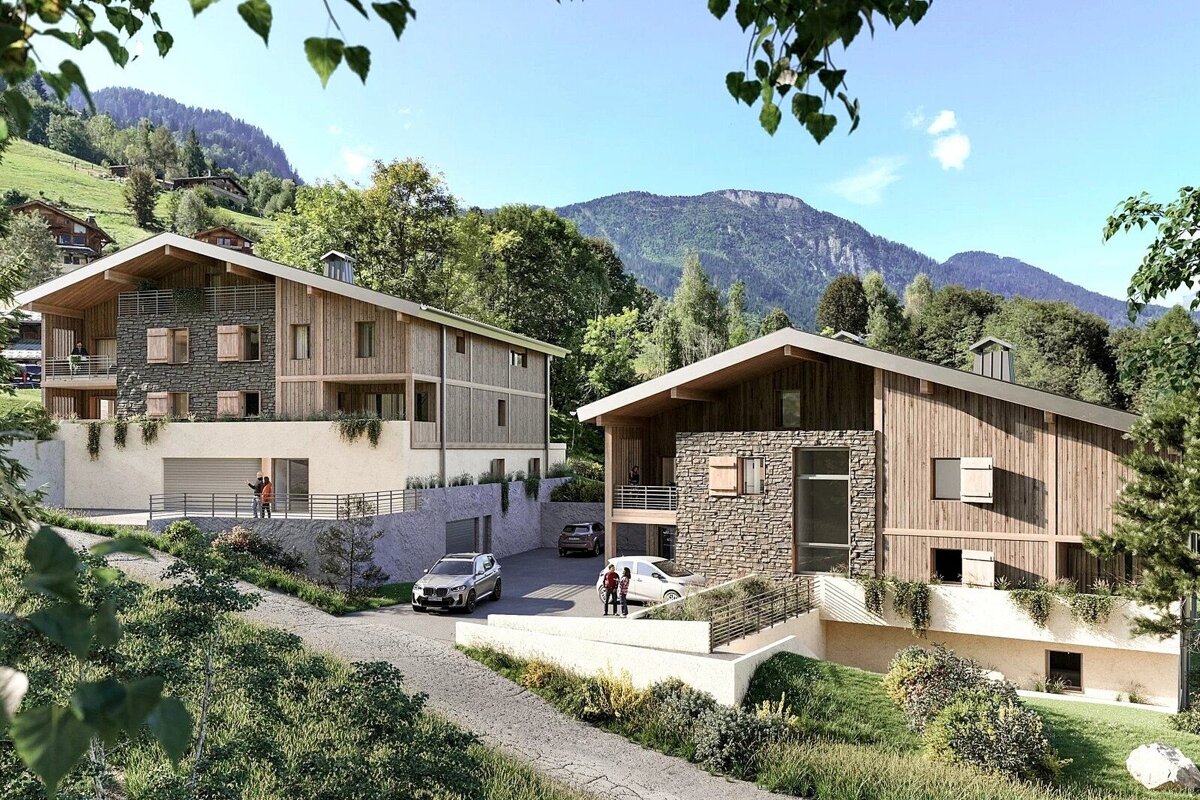 Two modern chalets with wood and stone facades are nestled in a lush green mountain valley under a blue sky. Cars are parked outside.