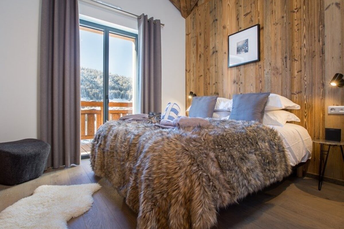 Cozy cabin bedroom with a faux-fur blanket on the bed, wood-paneled walls, and glass doors opening to a snowy mountain view.