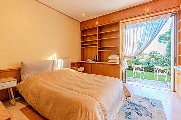 A sunny bedroom with a comfy bed, built-in wooden storage, and a sliding door to a green garden patio with white chairs.