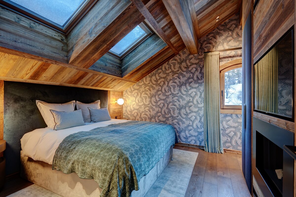 Cozy attic bedroom with rustic wooden beams, skylights, and a comfortable bed. Patterned wallpaper and a window with a snowy view.